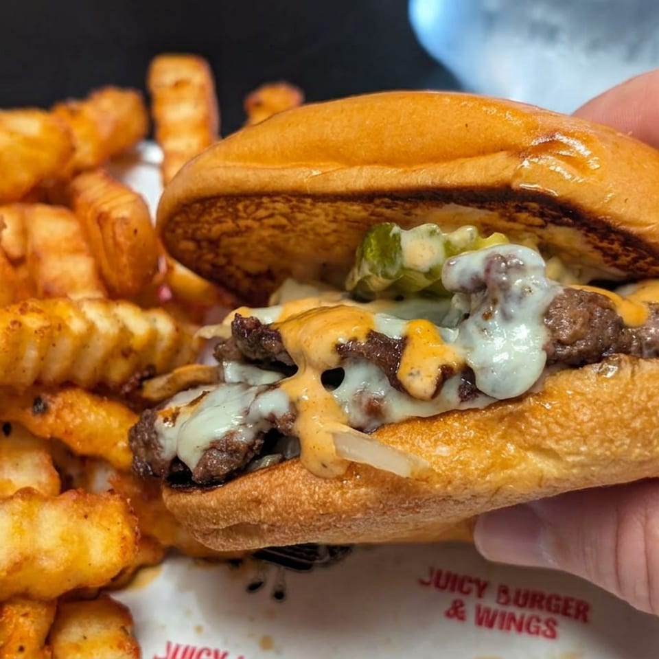 Hand holding a juicy cheeseburger with melted cheese and pickles beside seasoned crinkle-cut fries.